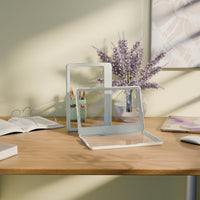 Two transparent stands on a wooden surface with a vase of flowers and books in the background.