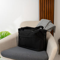 A black targus tote bag with top handles sits on a gray upholstered armchair in a modern interior setting. A folded gray blanket is draped over the back of the chair, and a wooden slatted wall panel is visible behind it.