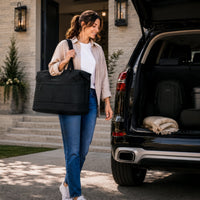 A woman carrying a large black duffel bag walks beside an open black SUV trunk in a driveway. The trunk contains a matching black backpack and a folded beige blanket.