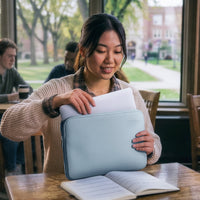 A woman in a beige knit sweater smiles while placing a white document into a light blue laptop sleeve on a wooden table in a cafe setting with a campus view in the background.