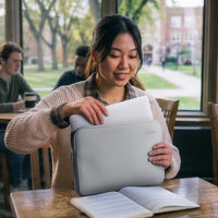 A woman in a beige knit sweater smiles while placing papers into a light gray laptop sleeve at a wooden table in a cafe, with a blurred campus background visible through the window.
