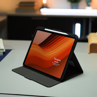 A black tablet case holding an iPad in landscape orientation on a light gray tabletop, with the screen on and showing the Targus logo. The background is a softly blurred office or home environment.