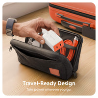 A dark gray zippered travel pouch on a wooden surface, shown open with a hand placing a white and orange power adapter inside. An orange suitcase is visible in the background, and the text 'Travel-Ready Design' appears at the bottom.