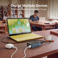 A portable multi-device charger sits on a wooden desk in a library or study, charging a yellow laptop, a smartphone, and other devices. The text 'Charge Multiple Devices' is displayed at the top, with students working in the background.