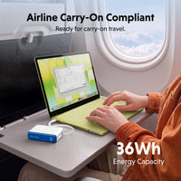 A portable power bank in white and blue is shown connected to a light green laptop on an airplane tray table, with a person typing on the keyboard. Text overlays read 'Airline Carry-On Compliant' and '36Wh Energy Capacity'.