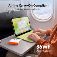 A portable white and orange power bank connected to a light green laptop on an airplane tray table, with a person typing. Text overlays read 'Airline Carry-On Compliant' and '36Wh Energy Capacity'.