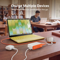A portable power station with orange and white accents is shown on a wooden desk in a library, charging a yellow laptop, a smartphone, and a small white device. White text at the top reads 'Charge Multiple Devices.