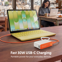 A portable white and orange USB-C power bank connected to a yellow laptop on a wooden table, with a woman working in a cafe in the background. Text overlay reads 'Fast 30W USB-C Charging'.