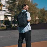 Man with a black backpack standing on a basketball court with trees and buildings in the background