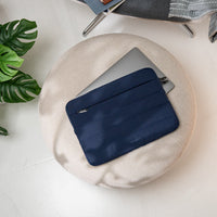 Navy laptop sleeve on a beige cushion with a plant and chair in the background