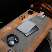A light blue laptop sleeve with a small silver logo, resting on top of a closed silver laptop, set on a gray felt desk mat on a wooden desk.