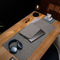 A gray laptop sleeve with a zippered closure sits on top of a closed silver laptop on a gray desk mat, arranged on a wooden desk with other lifestyle props including blue over-ear headphones, sunglasses, a black table lamp, and a white instant camera.