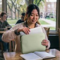 A woman in a cozy cafe setting holds a sage green laptop sleeve containing a white laptop. She is wearing a beige knit sweater, with blurred students and a campus background visible through the window behind her.