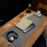 A light sage green laptop sleeve with a zipper closure, shown on a gray felt desk mat on a wooden desk. The scene is styled with a laptop inside the sleeve, blue over-ear headphones, sunglasses, a black table lamp, and a white instant camera on a blue notebook.