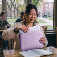 A woman in a beige knit sweater smiles while holding a lavender laptop sleeve with a white laptop inside, set in a bright cafe or study with a window showing a blurred campus background.