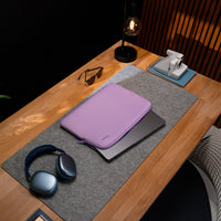 A lavender laptop sleeve with a matching zippered pouch rests on top of a closed silver laptop, placed on a gray felt desk mat on a wooden desk. A pair of blue wireless headphones, sunglasses, a black desk lamp, and a white instant camera are also visible in the scene.