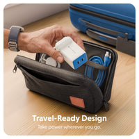 A dark gray zippered travel pouch on a wooden surface, shown with a hand placing a white and blue wall charger into its mesh pocket. A blue suitcase is blurred in the background, and the text 'Travel-Ready Design' is displayed at the bottom.