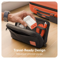 A hand inserts a white and orange power adapter into an open dark gray zippered travel pouch containing an orange cable, on a wooden surface with an orange suitcase in the background. Text at the bottom reads 'Travel-Ready Design' and 'Take power wherever you go.