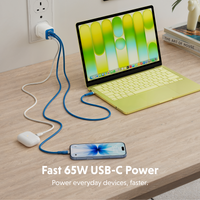 A 65W USB-C power cable setup on a light wood desk, with a laptop, phone, and earbud charging dock plugged in. The cables are blue and white, and the scene includes text reading 'Fast 65W USB-C Power' at the bottom.