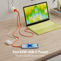A 65W USB-C power cable set shown on a wooden desk, with one orange cable and one white cable. The orange cable is plugged into a laptop with a light green keyboard cover, and the white cable is plugged into a white earbud case.