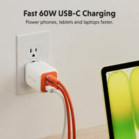 A white and orange USB-C power adapter plugged into a wall outlet, with an orange cable and a white cable plugged into it. Text at the top reads 'Fast 60W USB-C Charging' and a tablet is partially visible on the right.