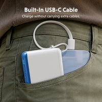 A close-up of a white portable power bank with a built-in white USB-C cable, tucked into the back pocket of olive green pants. A blue smartphone is partially visible behind it.