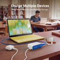 A portable multi-device charger sits on a wooden desk in a library, connected to a yellow laptop, a smartphone, and other devices. The charger has a white and blue design with the brand name 'EAGLE' visible on the side.