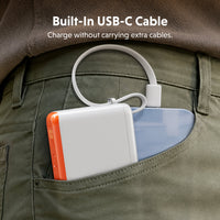 A close-up of a white portable power bank with an orange side edge and a built-in loop cable, tucked into the back pocket of olive green pants. A smartphone is partially visible behind it.