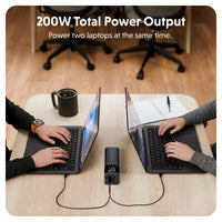 A portable black power station connected to two laptops on a light wood desk in an office setting, with text at the top reading '200W Total Power Output' and 'Power two laptops at the same time.'.