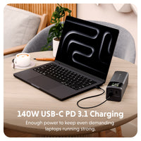 A laptop sits on a light wood table connected to a black 140W USB-C PD 3.1 power charger.