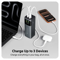 A portable black power bank with multiple USB-C ports is shown charging a laptop, a smartphone, and a white earbud case on a light gray background. Text at the bottom reads 'Charge Up to 3 Devices' and 'Charge everything in your bag, all at once.