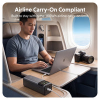 A man seated in an airplane cabin uses a laptop on a fold-out tray table while a dark gray portable power bank sits on the table in the foreground. Text at the top reads 'Airline Carry-On Compliant' and 'Built to stay within the 100Wh airline carry-on limit.