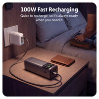 A portable gray power bank with a digital display is shown on a bedside table, connected to a wall charger and a brown leather wallet, with a person packing a suitcase in the background on a bed. Text at the top reads '100W Fast Recharging'.